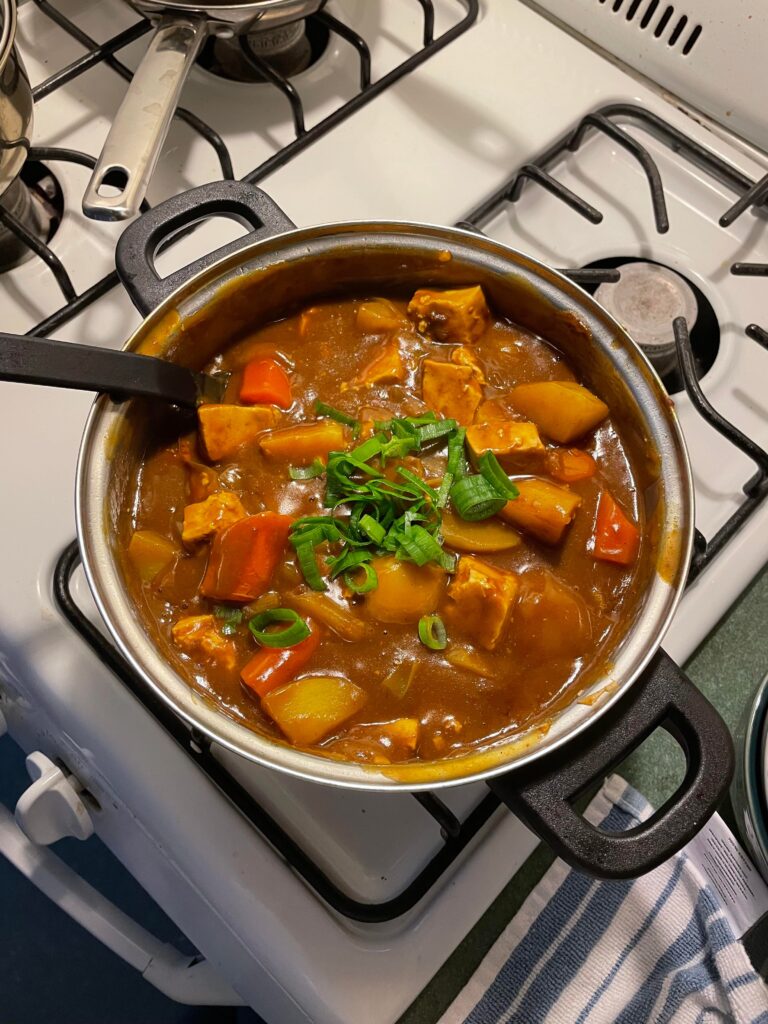Ashley's Japanese curry is placed on the stove and garnished with green onions. There are visible bits of potatoes, tofu, carrots, and onions chopped up. The curry is simmering.